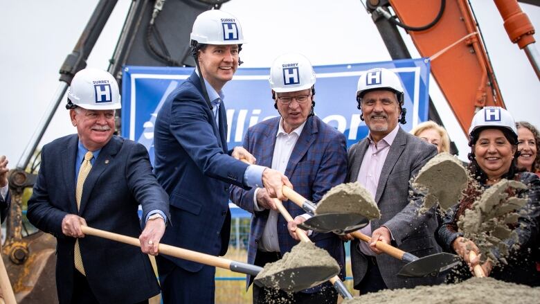 A group of people in white hard hats with the letter H on them toss shovels full of sand on a pile.