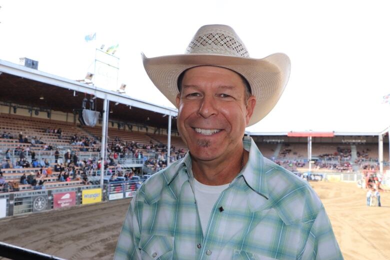 A man wearing plaid and a cowboy hat smiles at the camera standing in front of a rodeo stadium. 