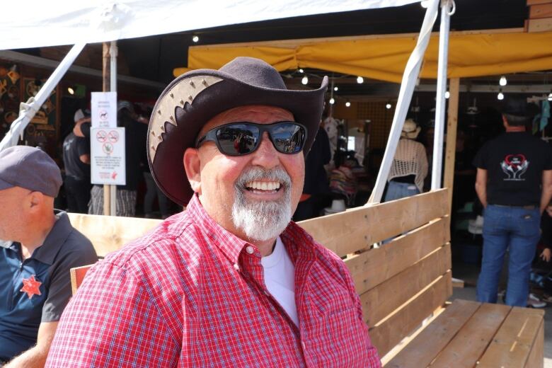 A man wearing a red plaid shirt and a cowboy hat smiles at the camera