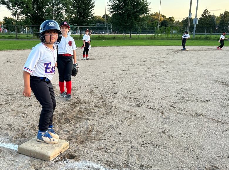 In the foreground, a girl stands poised on third base. In the background, players from the fielding team stand.