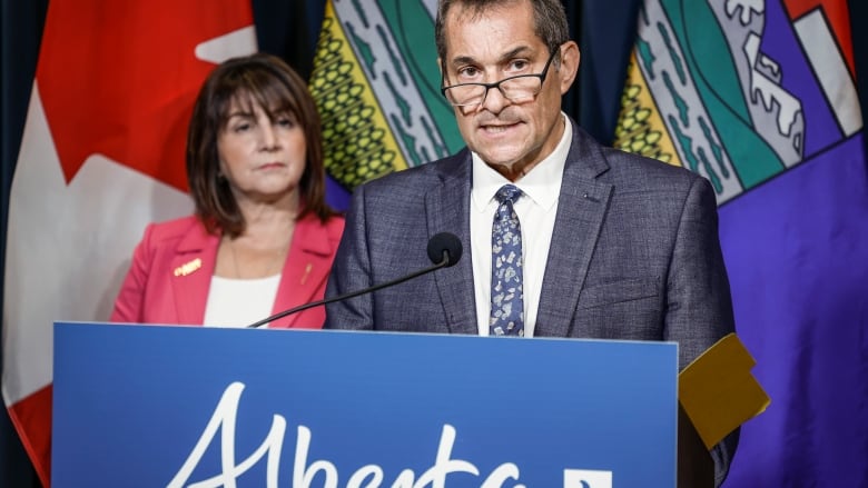 a bespectacled man speaks at a lectern while a woman looks on. Canadian and Alberta flags in the backdrop.