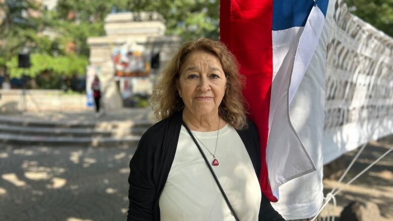 A woman stands outside at a park and looks into the camera. She stands beside a tree trunk from which hangs a Chilean flag. There's also a banner connecting the tree trunk to another off-frame with photos from different people who were killed or went missing during the dictatorship. 