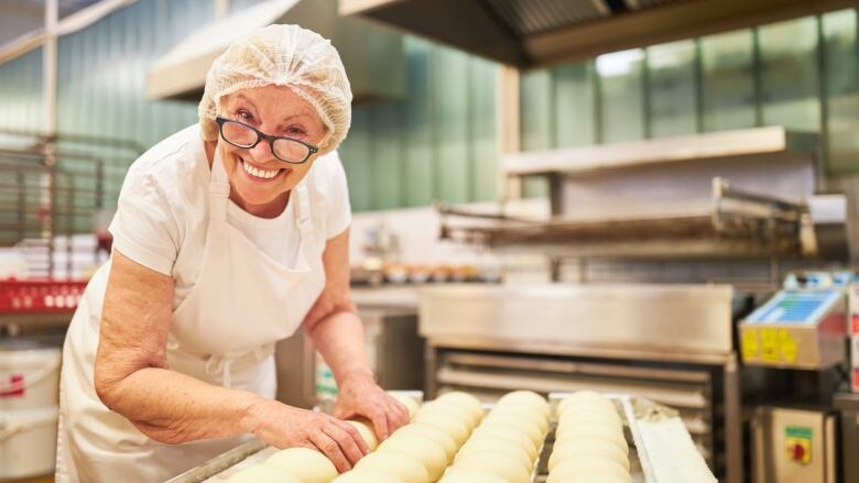 An older woman baker smiles as she handles some buns. 