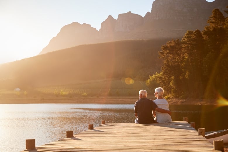 A senior couple sits on a wooden jetty by a lake.