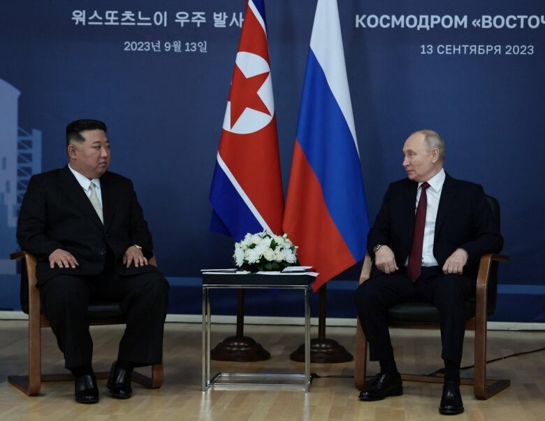 Two men sit in chairs, each wearing a suit and tie, in front of a backdrop featuring a pair of flags.