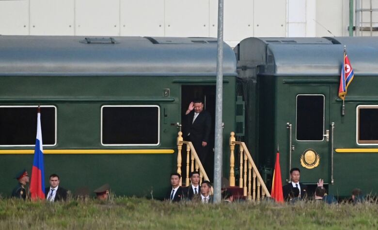 One men stands in a doorway of a train as several other men are shown on the ground in front of the train.