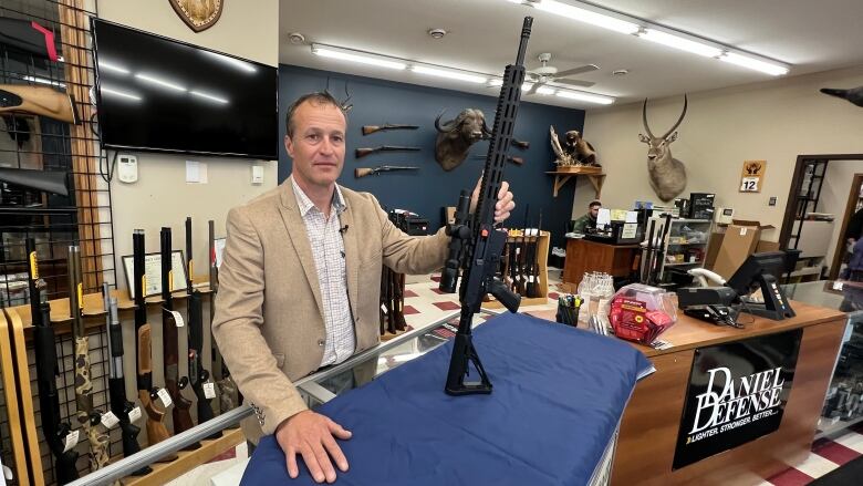 A man in a checkered shirt and tan suit jacket stands in front of a display of long-barreled guns