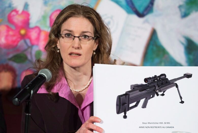 A woman in a lavender-coloured collared shirt and black suit jacked holds a photograph of an assault weapon.