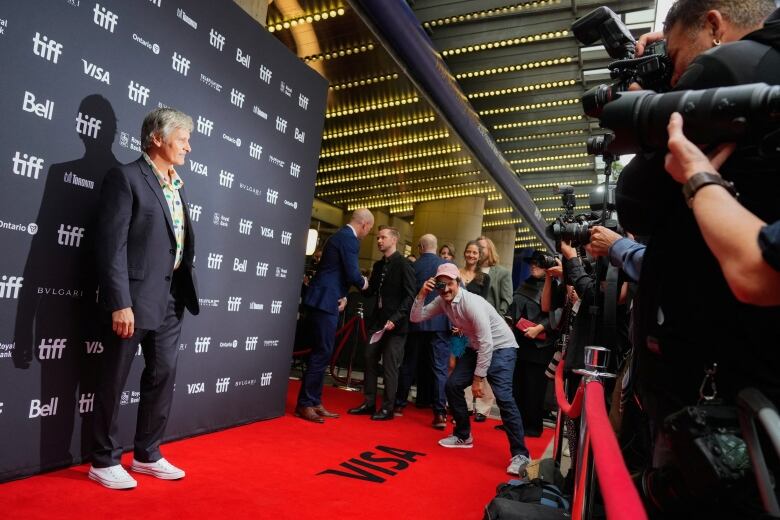 Film director faces a phalanx of photographers on the red carpet at TIFF, with another person taking photo in the photographer's direction.