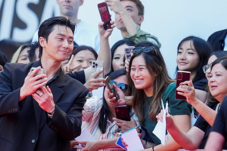 Asian actor in black jacket holds a cellphone taking photos with fans during TIFF>