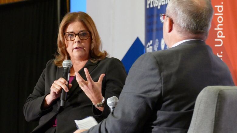 A woman holding a microphone speaks to a man who is facing away from the camera. 