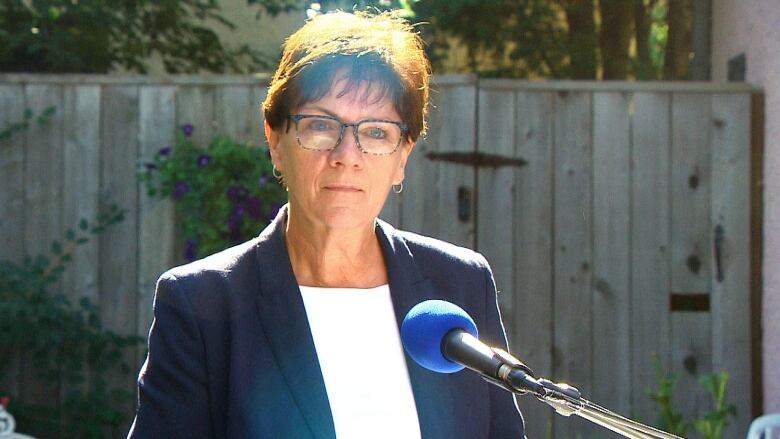 A woman in a blazer and white shirt stands behind a podium with a microphone.