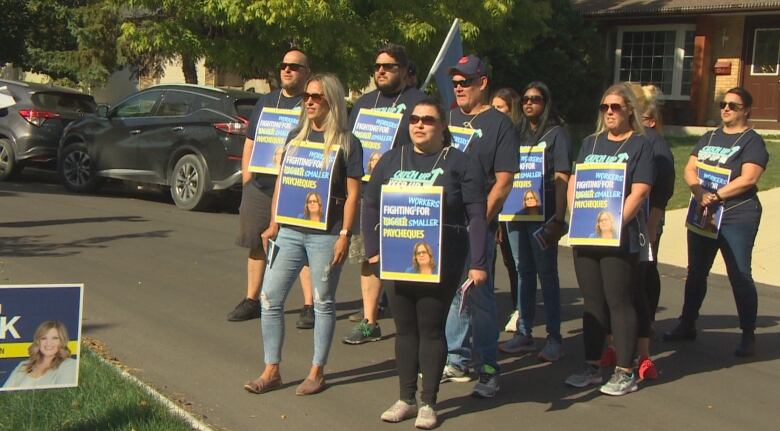 Striking protesters stand on the street, wearing matching black shirts and carrying signs.