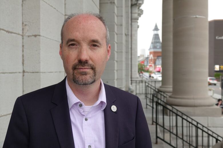 A man with a goatee and salt and pepper hair stands next to a grey brick wall. Behind him are several large pillars.