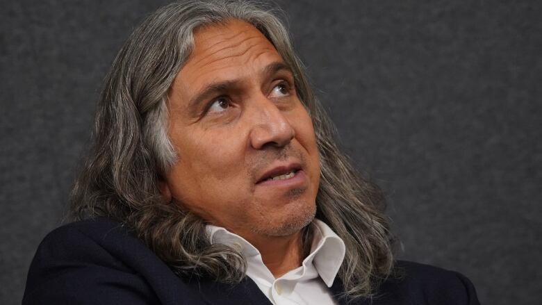 Close up shot of a man's face with long grey hair in a suit, looking up. 