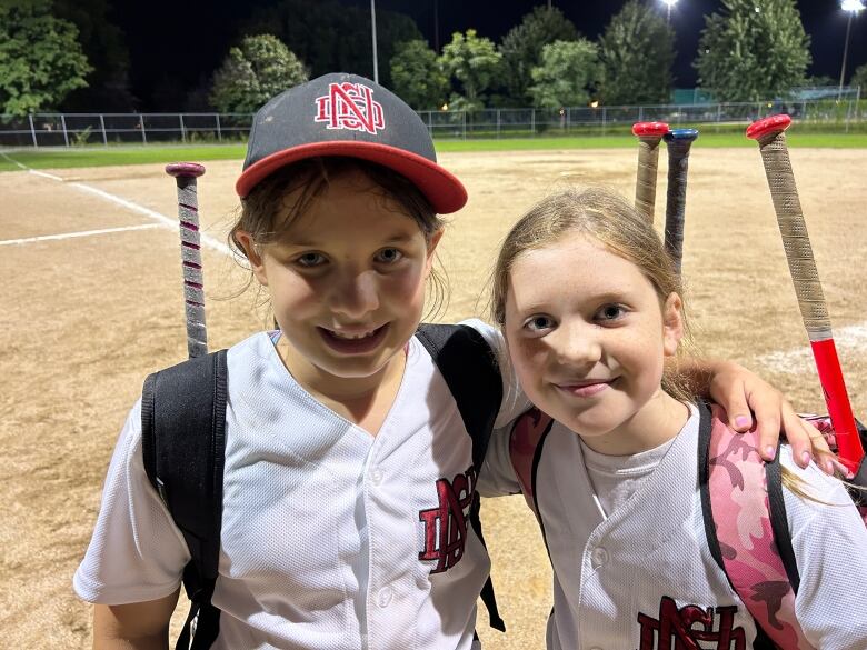 Two preteen girls in baseball gear with their arms around each other's shoulder smile into the camera.