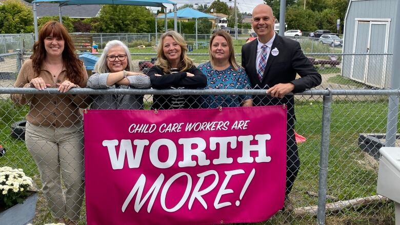 Five people standing behind a fence with a sign that says, 'Child care workers are worth more.'