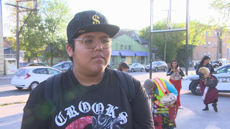 A person wearing a black t-shirt and black cap stands in front of a street in a parking lot. Some people stand in the background.