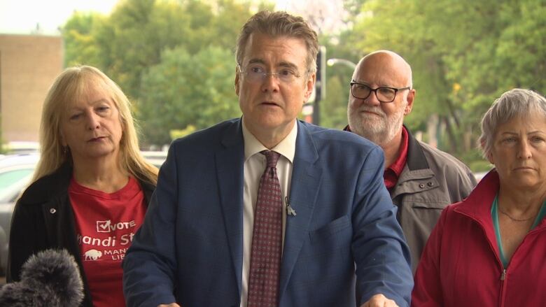 A man in a suit is speaking in front of microphones with people standing behind him.