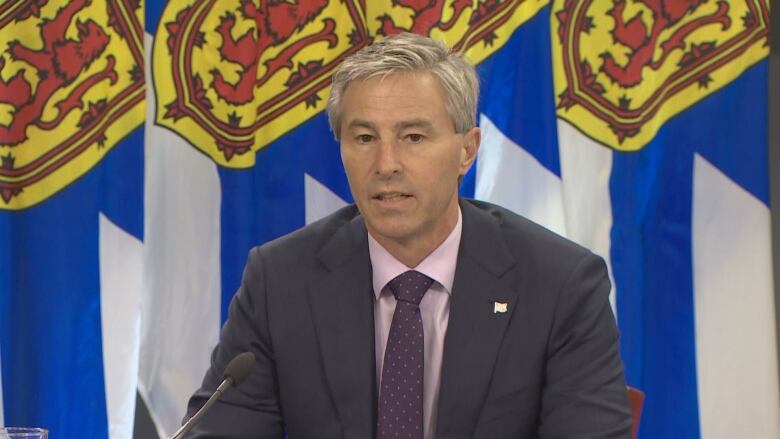 A man in a dark suit, pink shirt and purple tie sits in front of flags.