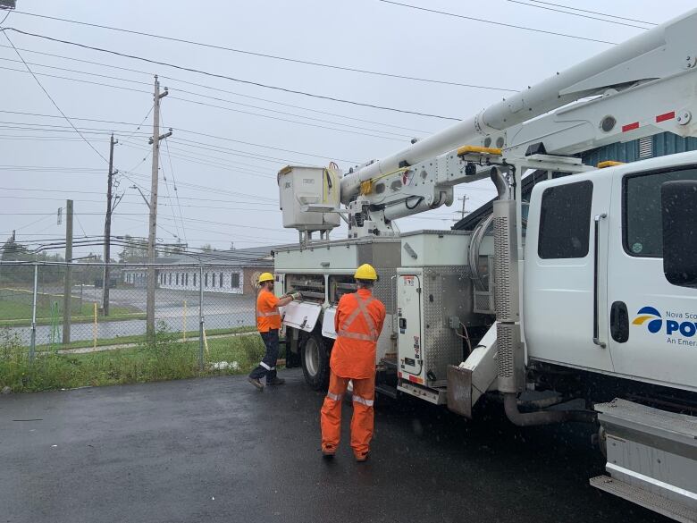 Two people wearing orange jumpsuits and yellow hard hats are seen next to a large white utility truck on a foggy day.