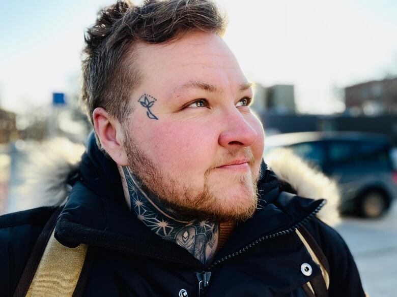 A man with short brown hair and tattoos on his neck and temple looks off into the distance.