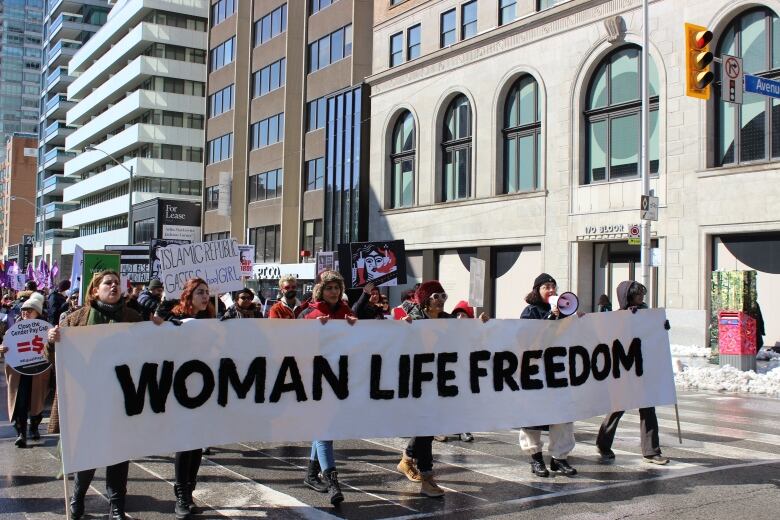 A crowd of people walk a city street with a banner in front of them, which says Woman Life Freedom