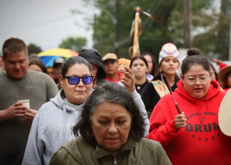 A woman in grey shirt and sunglasses raises a small candle while walking among a large crowd of people.