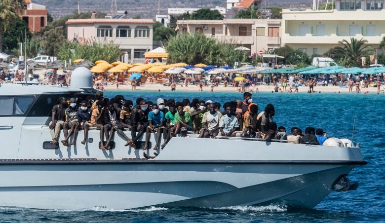 A large boat filled with people on its deck travels through the water, with the buildings and shoreline of a beach town shown behind it.