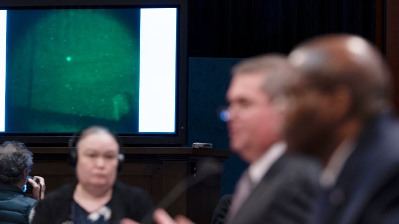 A TV screen shows a white blob on an otherwise dark sky as people in suits speak in a government hearing room.