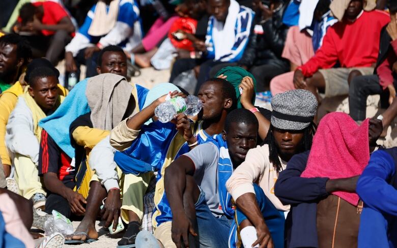 Dozens of people sit on the paved ground outside, in the sun, some with towels covering their heads and others drinking water.