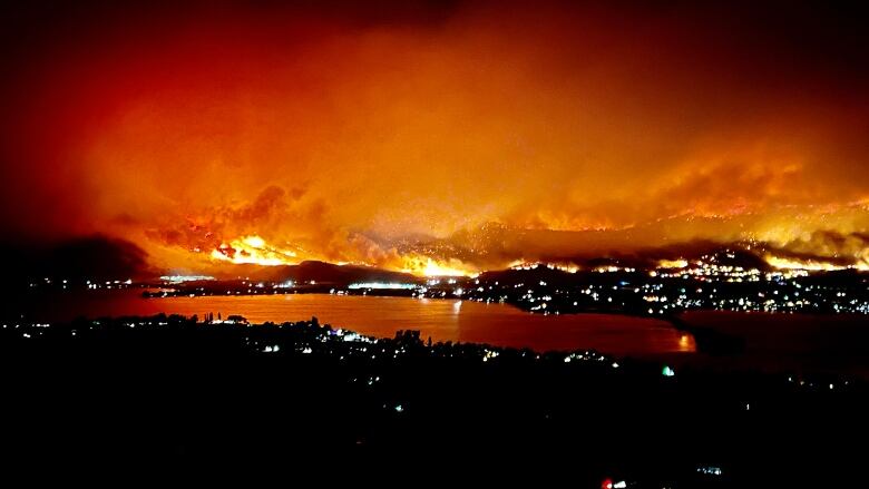 A massive, billowing fire is encroaching onto a town that's lit up with many with many lights on due to it being nighttime that has a body of water in front of it that's reflecting the orange glow. 