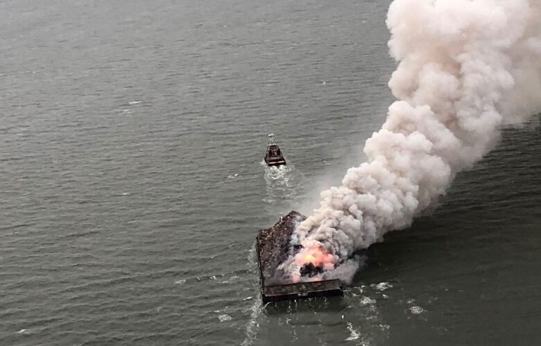 Thick white smoke billows from a barge of scrap metal on fire.