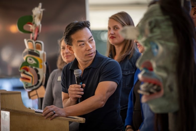 A man holding a microphone surrounded by others at a podium looks over his shoulder at an Indigenous mask carving.