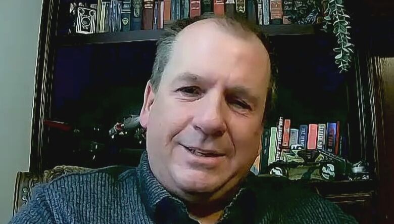 Man with light brown hair sits in front of a bookcase. 