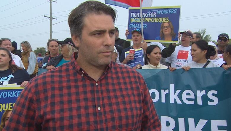 A man is seen looking to his left. Behind him, a group of people are shown holding up signs.