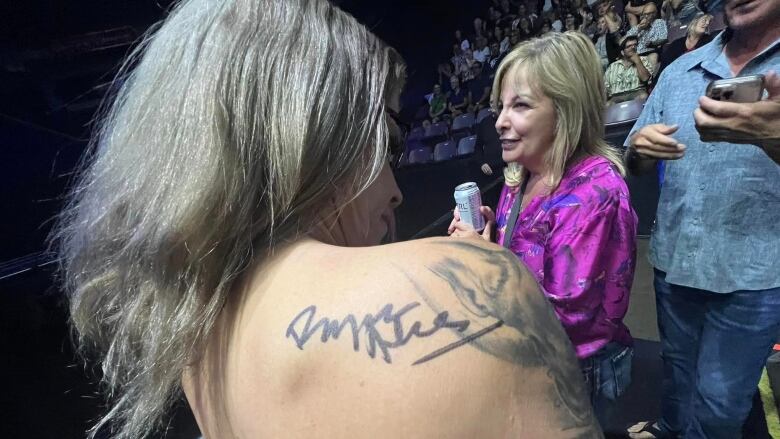 A woman shows a signature, in black felt marker, on the back of her right shoulder, while standing at the front of a rock concert.