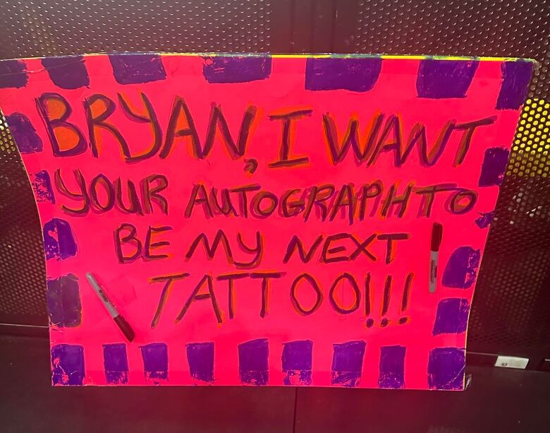 A sign reading, Bryan, I want your autograph to be my next tattoo, is shown.