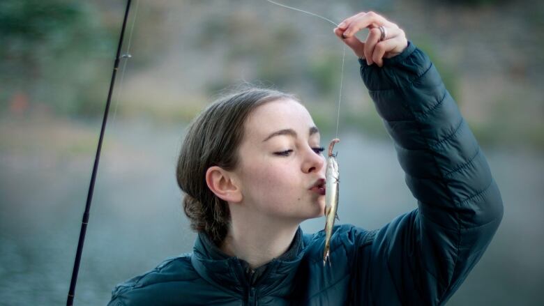 A teen girl wearing a black down jacket holds a small fish on a fishing line to her lips