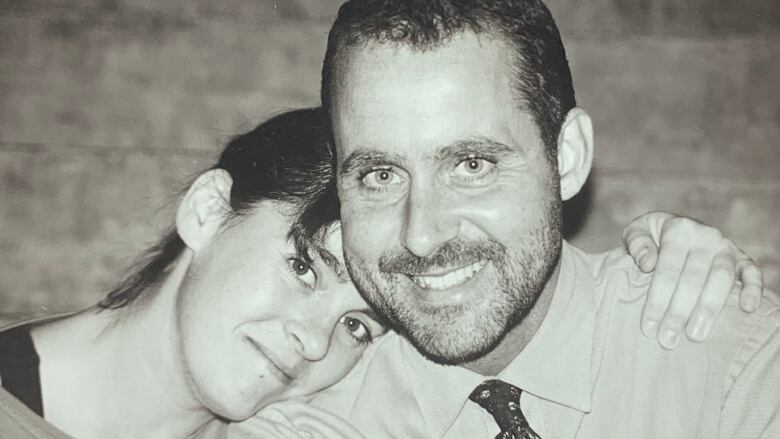 A woman rests her head on a man's shoulder as they both smile at the camera in a black and white photo.