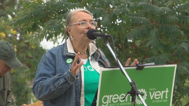 A woman wearing a denim jacket and green t-shirt speaks into a microphone behind a podium.