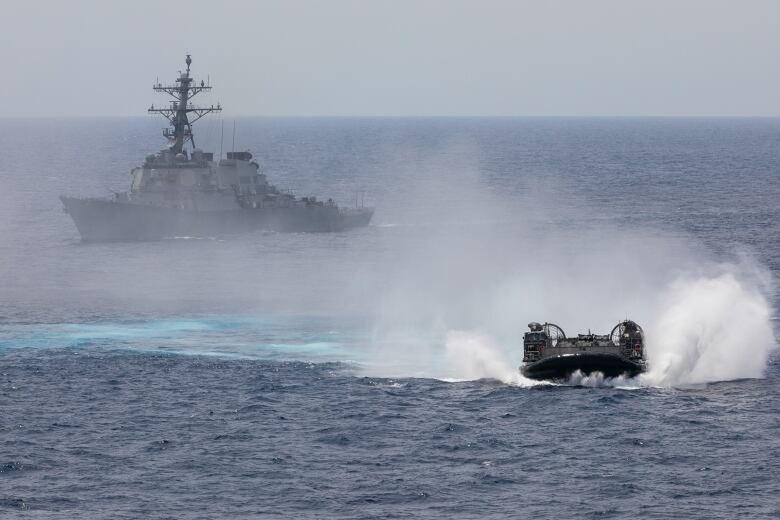 An amphibious vehicles navigates waters as a warship is seen in the background.