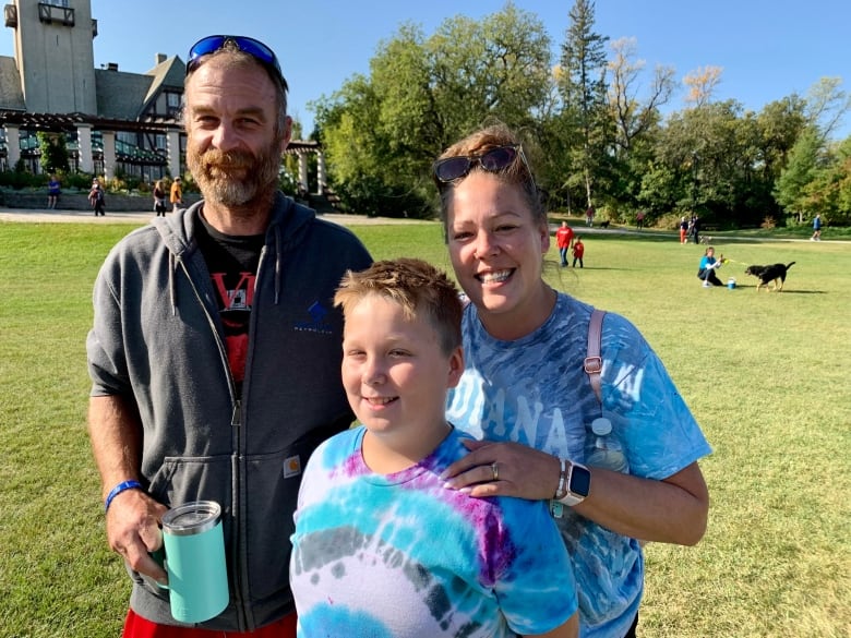 An 8 year old boy wearing a t-shirt stands in a park with his parents.