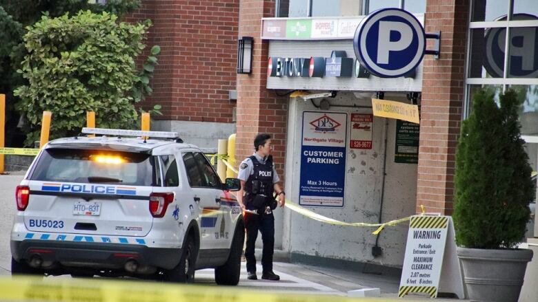 A parkade entrance is cordoned off by police tape and an officer stands outside it with an RCMP vehicle next to him.