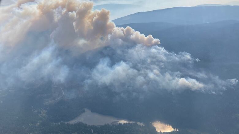 Smoke rises from a fire near a lake on a sunny day from above.