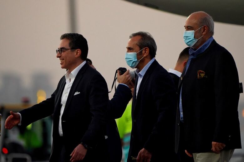 Three men in suits walking in a row. The man in front is smiling and extending his hand toward someone off camera. The two men behind him are wearing medical masks. 