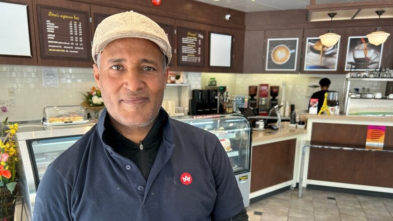 A cafe owner in a blue polo shirt and beige hats poses for a photo in his cafe.