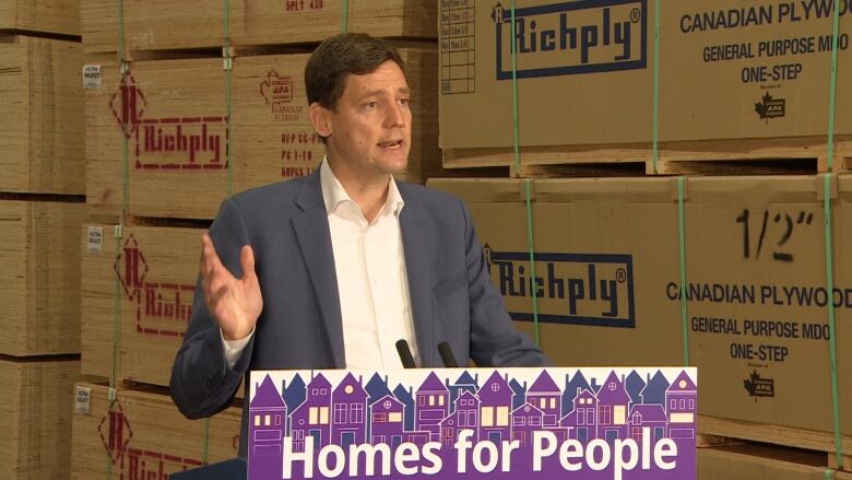 David Eby waves a hand while speaking a podium. Stacks of plywood are seen in the background.