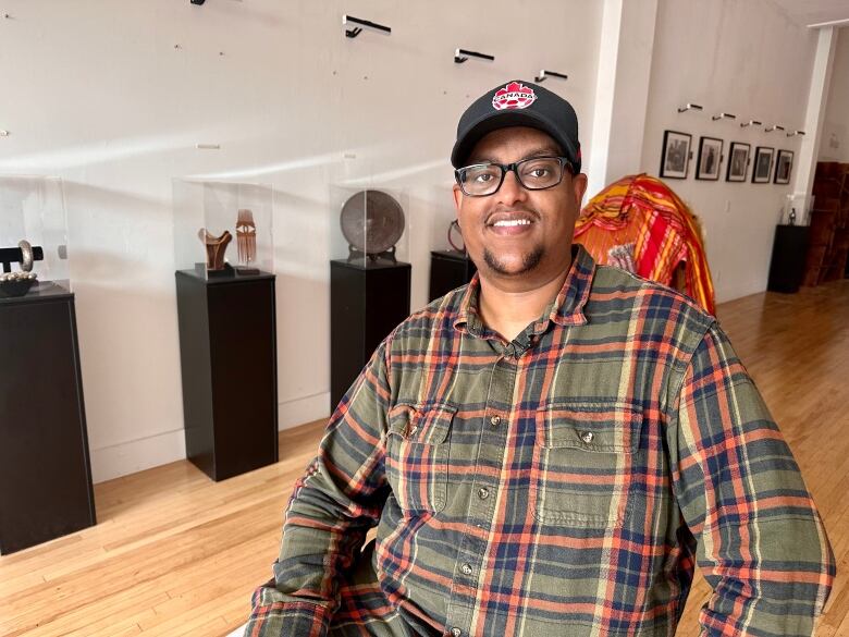 A man in flannel shirt, ballcap and glasses sits inside in front of artifacts in a glass case