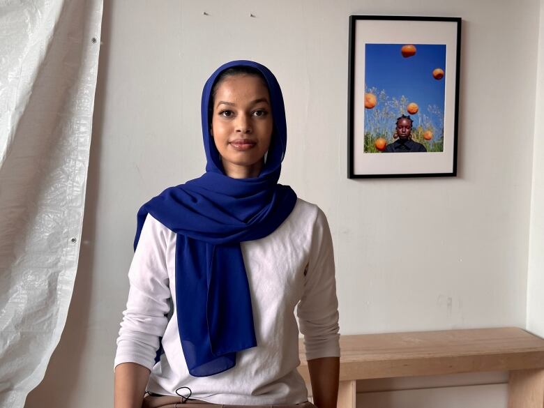 A young woman in a blue hijab sits inside in front of a painting of a woman and oranges.